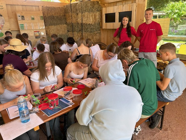 Bild: Das Bild zeigt Jugendlichen, die in einer Holzhütte an Tischen sitzen. Jeder hat ein Schraubglas mit Einzelteilen zum Zusammenbauen, für ein für ein Solarauto.